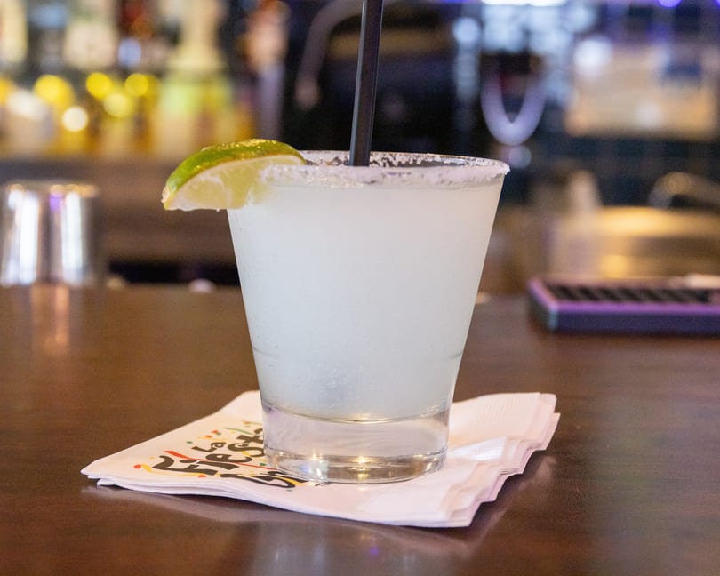 A frosty margarita with a salted rim and lime wedge, on a napkin at a bar.