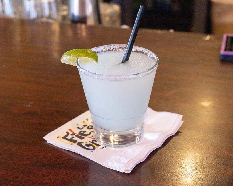Frozen margarita with a lime wedge and salted rim on a branded napkin at a bar.