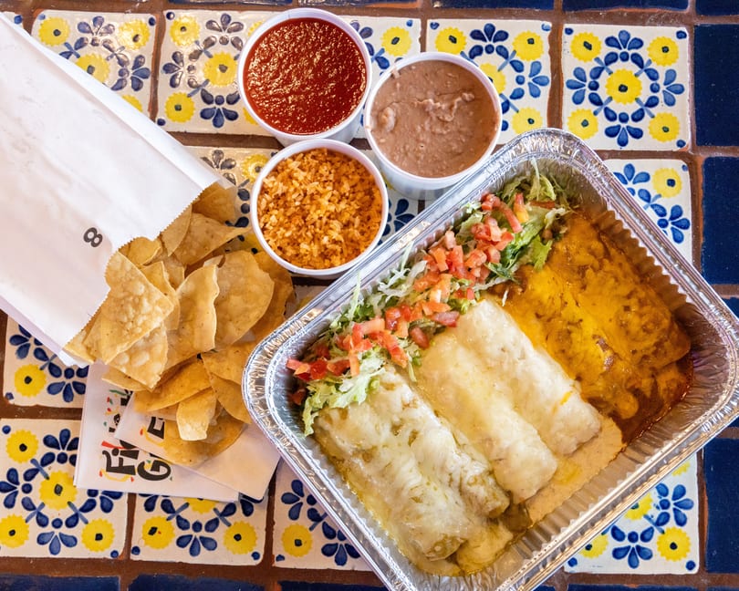 Mexican takeout meal with enchiladas, rice, beans, salsa, and tortilla chips on a tiled table.