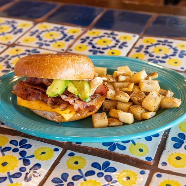 Breakfast sandwich with bacon, avocado, and cheese on a bagel, served with home fries on a blue plate.
