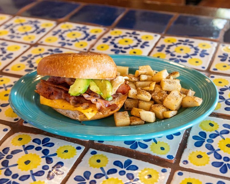 Breakfast sandwich with bacon, avocado, and cheese on a bagel, served with home fries on a blue plate.