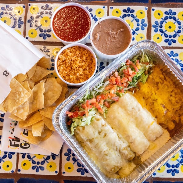 Mexican takeout meal with enchiladas, rice, beans, salsa, and chips on a colorful tiled table.