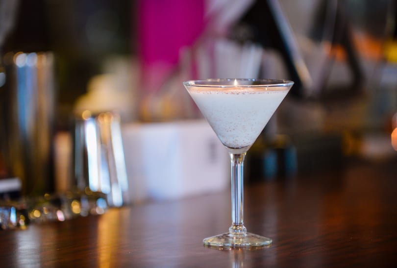 A creamy martini with a sprinkle of spices sits on a dark wooden bar.