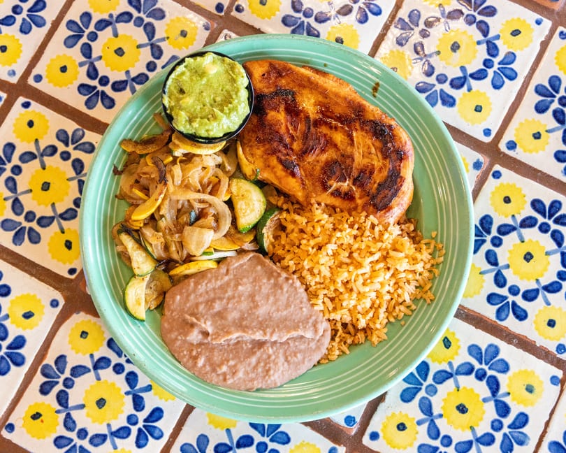 Grilled chicken, rice, refried beans, sauteed vegetables, and guacamole on a green plate.