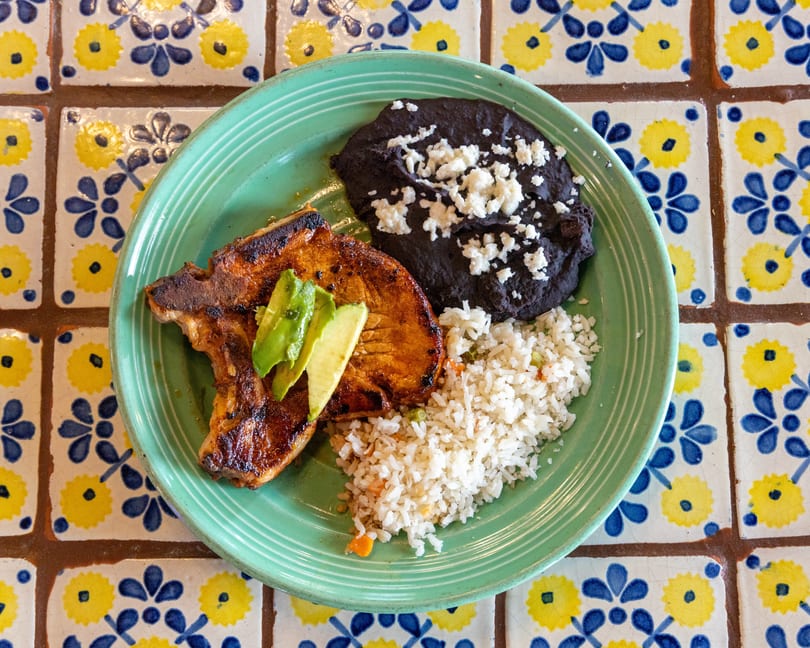 Grilled pork chop with avocado slices, black beans, and rice on a green plate over patterned tiles.