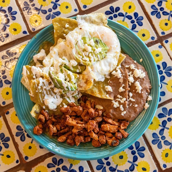 Chilaquiles with green sauce, fried eggs, avocado, refried beans, and chorizo on a blue plate.