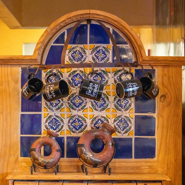Black "Fiesta Grande" mugs hang from a wooden rack with blue and yellow patterned tiles.