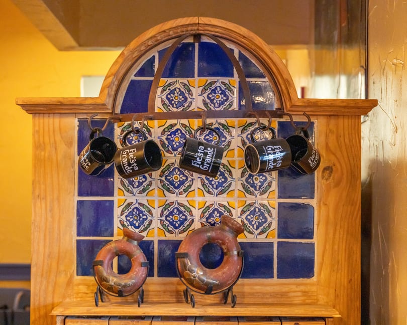 Black "Fiesta Grande" mugs hang from a wooden rack with blue and yellow patterned tiles.