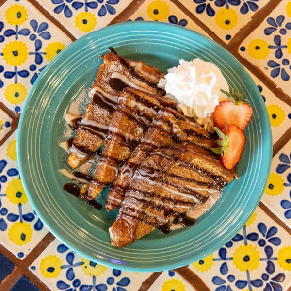 Churro French Toast on a teal plate, drizzled with chocolate and condensed milk, with whipped cream and strawberries.