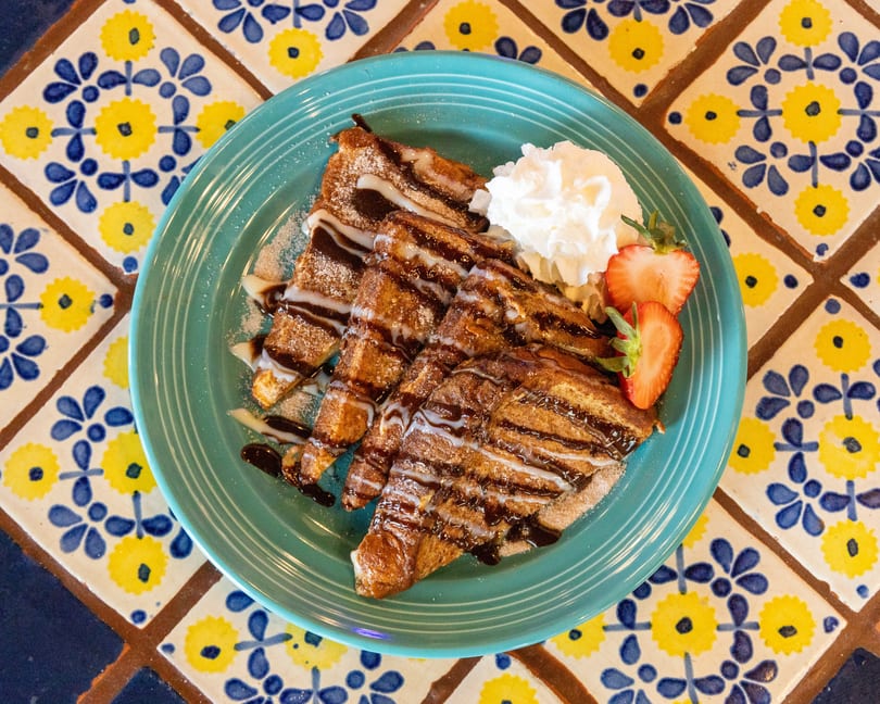 Churro French Toast on a teal plate, drizzled with chocolate and condensed milk, with whipped cream and strawberries.