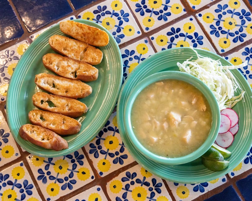 Pozole verde with shredded cabbage, radishes, limes, and toasted bread on a colorful tiled table.