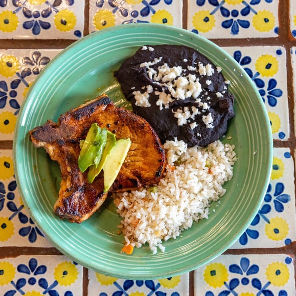 Grilled pork chop with avocado slices, black beans, and rice on a green plate over patterned tiles.