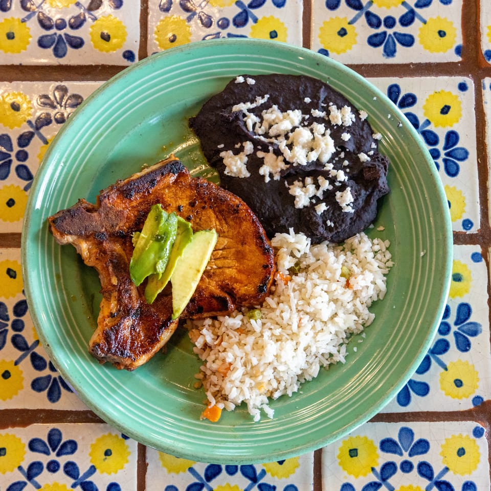 Grilled pork chop with avocado slices, black beans, and rice on a green plate over patterned tiles.