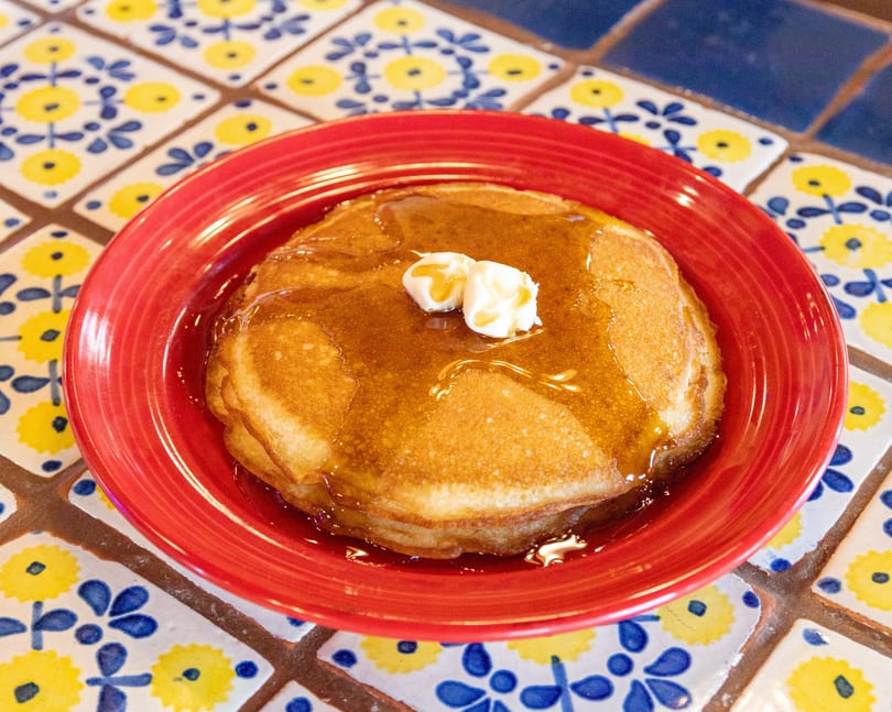 Golden pancakes with butter and syrup on a red plate, on a colorful tiled table.