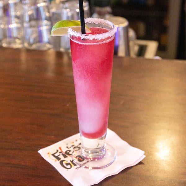 A tall glass of pink frozen margarita with a lime wedge and salt rim on a bar.