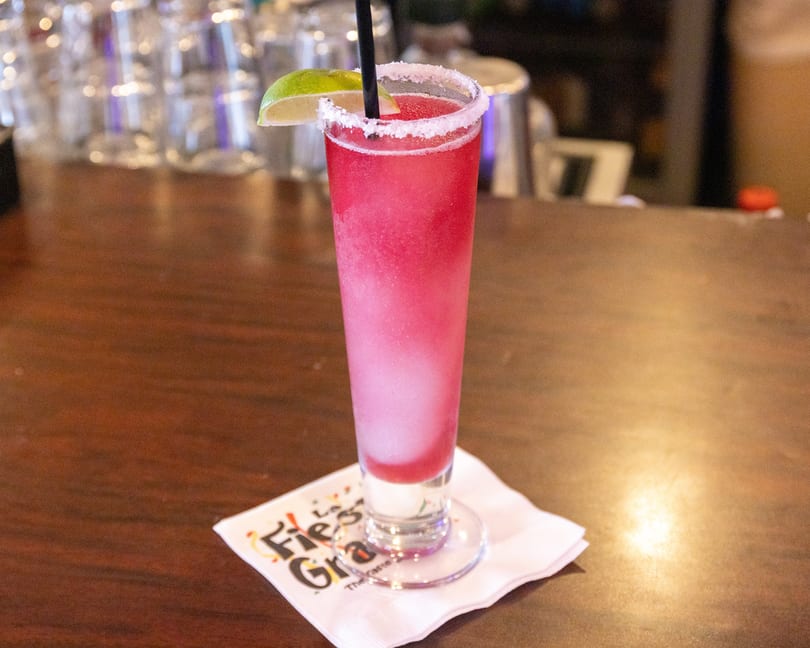 A tall glass of pink frozen margarita with a lime wedge and salt rim on a bar.
