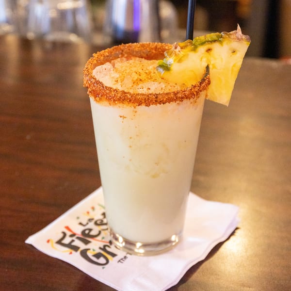 A creamy cocktail with a chili-lime rim, garnished with a pineapple slice and black straw.