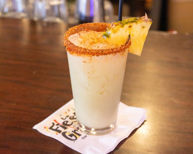 A creamy cocktail with a chili-lime rim, garnished with a pineapple slice and black straw.