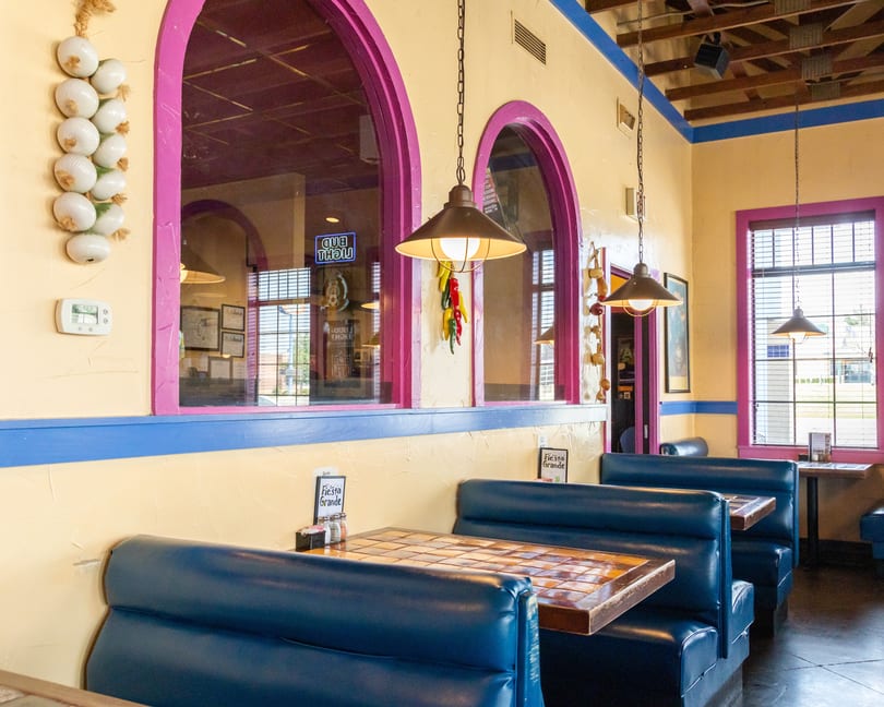 Interior of a vibrant Mexican restaurant with blue booths, tiled tables, and arched windows with pink trim.