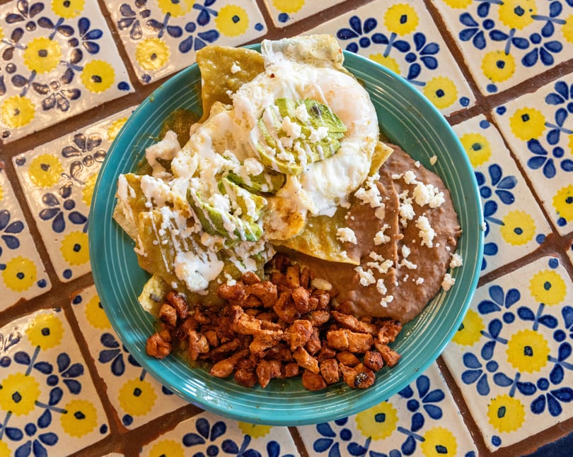 Chilaquiles with green sauce, fried eggs, avocado, refried beans, and diced chicken on a blue plate.