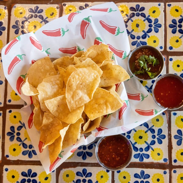A basket of tortilla chips with three small bowls of salsa on a colorful tiled table.