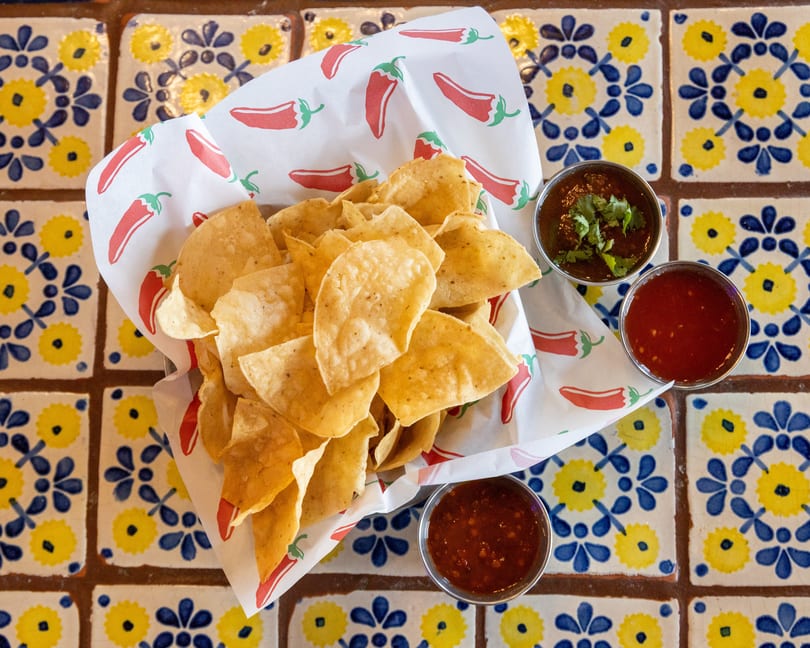A basket of tortilla chips with three small bowls of salsa on a colorful tiled table.