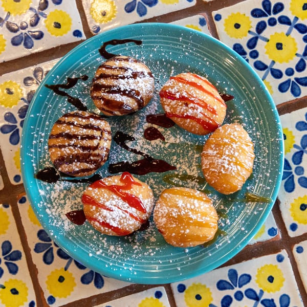 Six golden-fried dough balls drizzled with chocolate, strawberry, and caramel sauces, dusted with powdered sugar.