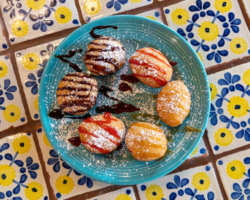 Six golden-fried dough balls drizzled with chocolate, strawberry, and caramel sauces, dusted with powdered sugar.