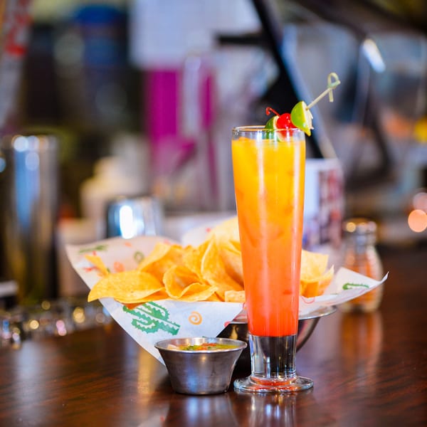 A tall, colorful cocktail with chips and salsa on a dark wooden bar.