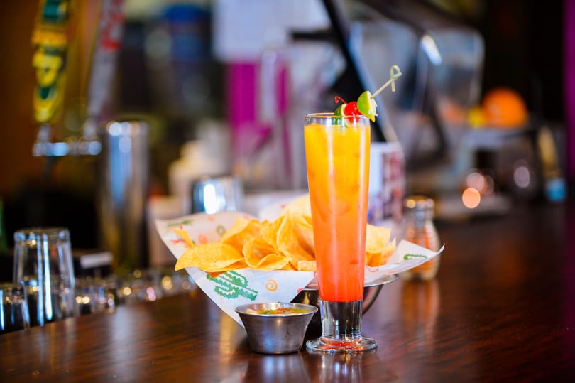 A tall, colorful cocktail with chips and salsa on a dark wooden bar.