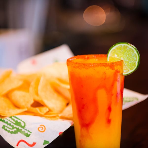 A vibrant orange and red drink with a lime slice next to a basket of tortilla chips.