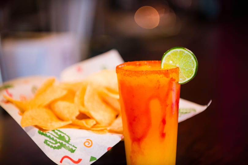 A vibrant orange and red drink with a lime slice next to a basket of tortilla chips.