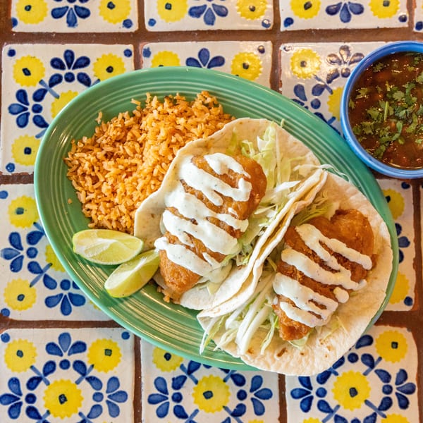 Two fish tacos with rice, lime wedges, and a side of beans on a colorful tiled table.