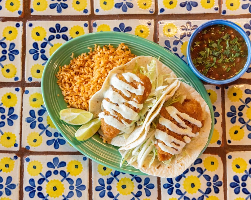 Two fish tacos with rice, lime wedges, and a side of beans on a colorful tiled table.