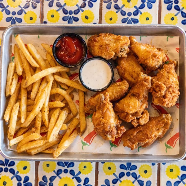 A metal tray of crispy fried chicken wings, french fries, ketchup, and ranch dressing on a colorful tiled table.