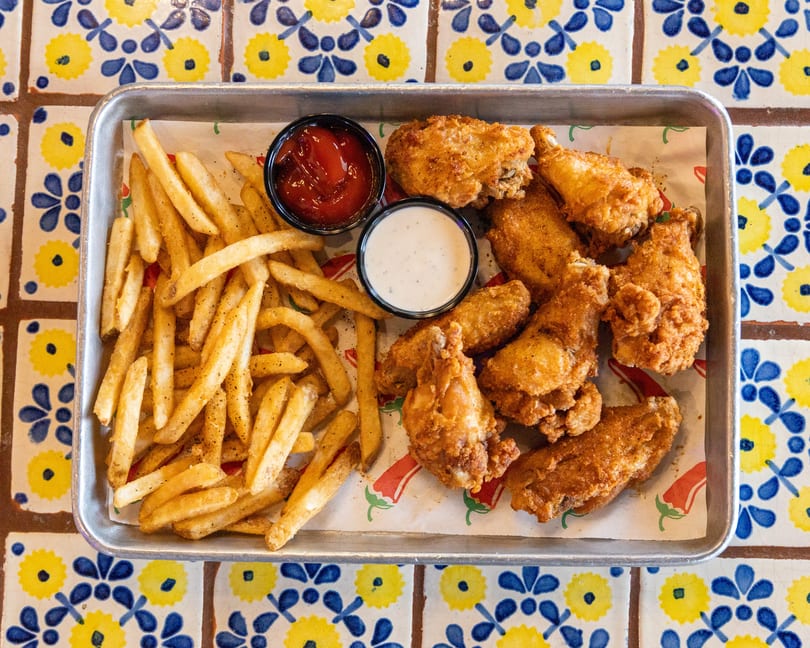 A metal tray of crispy fried chicken wings, french fries, ketchup, and ranch dressing on a colorful tiled table.