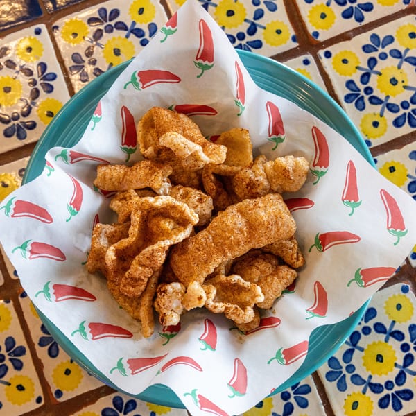 A teal bowl of seasoned chicharrones on chili pepper-patterned paper, set on a yellow and blue tiled table.