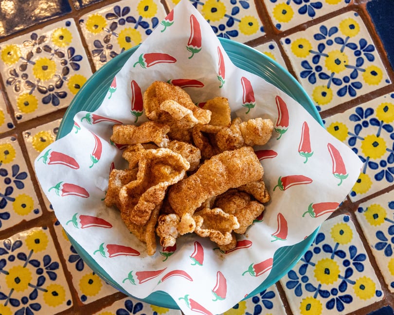 A teal bowl of seasoned chicharrones on chili pepper-patterned paper, set on a yellow and blue tiled table.