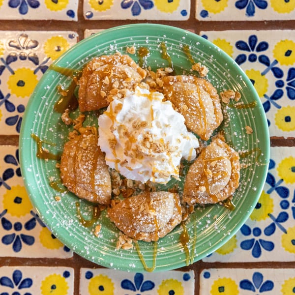 A green plate of golden-brown fried pastries with whipped cream, caramel, and powdered sugar on a tiled table.