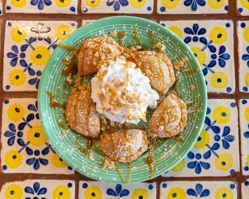 A green plate of golden-brown fried pastries with whipped cream, caramel, and powdered sugar on a tiled table.
