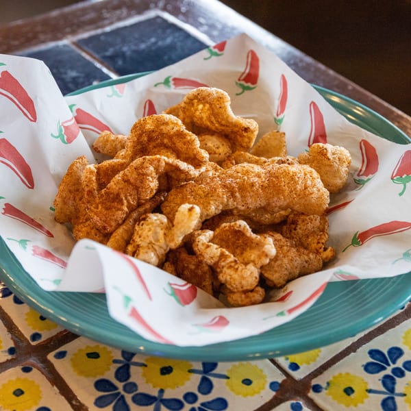 A teal bowl filled with crispy, seasoned chicharrones on chili-patterned paper, resting on a tiled table.