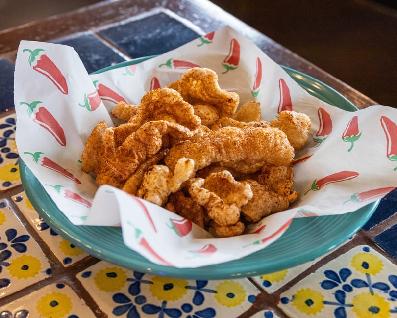 A teal bowl filled with crispy, seasoned chicharrones on chili-patterned paper, resting on a tiled table.