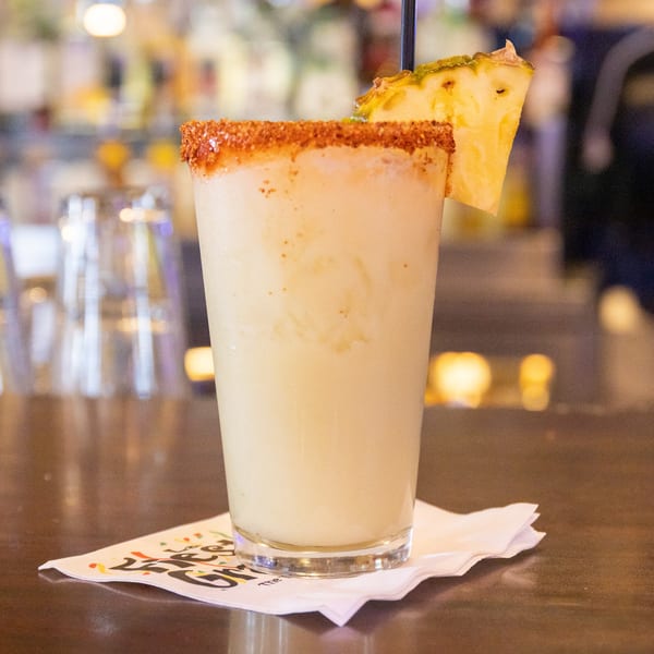 Frothy pineapple margarita with a chili-lime rim and pineapple wedge, on a bar with a branded napkin.