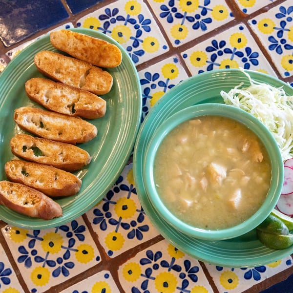 Pozole verde with shredded cabbage, radishes, and lime, served with toasted bread on a colorful tiled table.